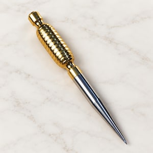 May include: A metal tool featuring a gold-colored, ribbed, cylindrical handle and a smooth, silver-colored, tapered conical point. The handle has a rounded top and connects to the pointed tip via a short, smooth gold-colored cylindrical section.