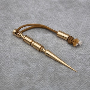 May include: A polished gold-tone metal plumb bob featuring a slender, pointed tip and a cylindrical body with decorative rings. A brown cord is attached to the top, ending in a matching gold-tone metal cylinder with a frayed, light-colored end.