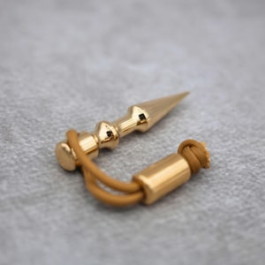 May include: A polished gold-toned metal plumb bob featuring a pointed tip and a cylindrical top section. A tan-colored cord is looped through the top of the plumb bob and connects to a small, matching gold-toned cylindrical weight at the end.
