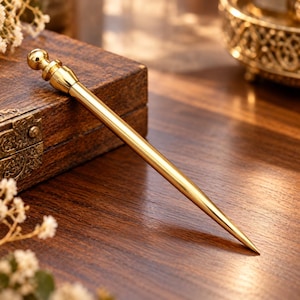 May include: A polished gold-toned metal hair stick featuring a smooth, tapered shaft and a decorative rounded finial top. The accessory has a reflective, metallic finish and a pointed end, designed with a simple, clean silhouette for hair styling.