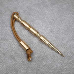 May include: A polished gold-tone metal spike tool featuring a cylindrical body with decorative ridges and a tapered point. A tan paracord lanyard is attached to the top, secured by a matching gold-tone metal bead at the end of the cord loop.