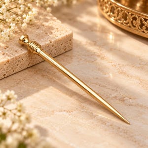 Könnte beinhalten: Ein schlanker Haarpick aus poliertem, goldfarbenem Metall mit einem glatten, spitz zulaufenden Schaft und einem dekorativen, gerippten zylindrischen Kopf. Das Accessoire hat eine reflektierende Metalloberfläche und ein spitzes Ende zum Fixieren.
