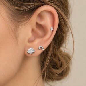 May include: A set of three silver-toned stud earrings featuring a cloud shape encrusted with clear stones, a small rainbow arch with clear stones, and a lightning bolt shape with a single clear stone, all worn in the earlobe and cartilage.