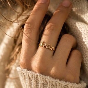 Op de afbeelding: Een goudkleurige metalen ring met de naam Ivette in een cursief, handgeschreven lettertype. De ringband is dun en gepolijst, waardoor de naam duidelijk op de vinger zichtbaar is met een gladde, reflecterende metalen afwerking.