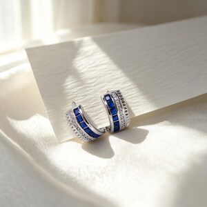 May include: A pair of silver-toned hoop earrings featuring a row of square-cut blue gemstones flanked by two rows of small, clear, round-cut stones. The earrings have a curved, wide band design with a polished metallic finish and secure closures.