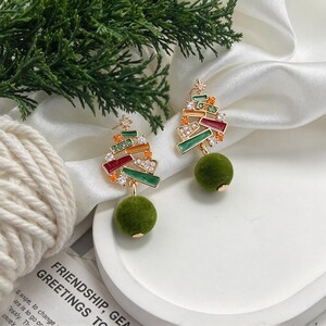 Könnte beinhalten: Ein Paar goldfarbene Ohrringe in Form von Weihnachtsbäumen mit geometrischen Emaille-Segmenten in Rot, Grün und Orange, akzentuiert mit klaren Kristallen und einem Stern, abgerundet mit einem hängenden grünen Samt-Pompon.