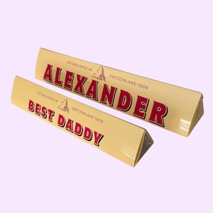 May include: Two triangular cream-coloured chocolate bar boxes with red lettering. The larger box reads ALEXANDER, ESTABLISHED IN SWITZERLAND 1908. The smaller box reads BEST DADDY, ESTABLISHED IN SWITZERLAND 1908. Both feature a small mountain logo.
