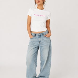 Könnte beinhalten: Ein weißes kurzärmeliges T-Shirt mit dem rosa Schriftzug "Fluent in yappanese" auf der Brust, kombiniert mit einer hellblauen Jeans mit weitem Bein, klassischem Knopf- und Reißverschluss sowie weißen Sportschuhen.