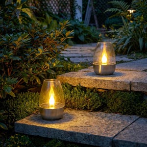 May include: Two outdoor lanterns with a clear glass shade and a metallic silver base. Each lantern contains a warm yellow LED light shaped like a flickering flame, providing illumination on stone steps surrounded by green garden foliage.