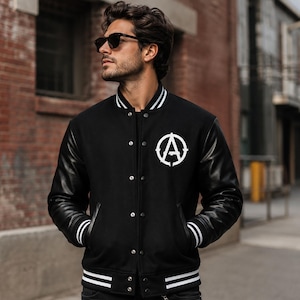 May include: A black varsity jacket featuring black leather sleeves, a snap-button front closure, and white striped ribbed trim on the collar, cuffs, and hem. A white circular logo with a stylized letter A is printed on the left chest area.