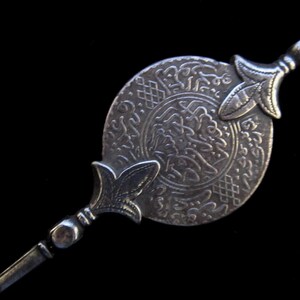 May include: A silver-toned decorative pin with an ornate circular design featuring Arabic script. The pin has two decorative points on either end.
