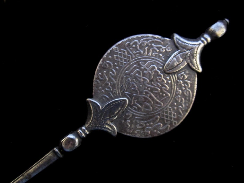 May include: A silver-toned decorative pin with an ornate circular design featuring Arabic script. The pin has two decorative points on either end.