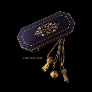 May include: A dark blue jewelry box with gold filigree and three gold necklaces with pendants. The pendants include a hand, a sphere with stars, and a hand with a green stone. The background is black.