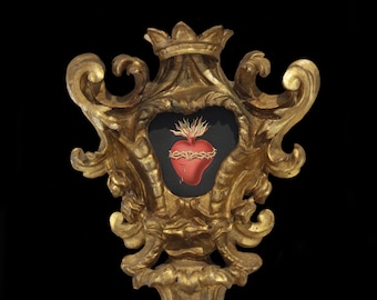 May include: A gold-painted wooden frame with ornate details, featuring a black and red image of a heart pierced with thorns and flames. The heart is surrounded by a black background.