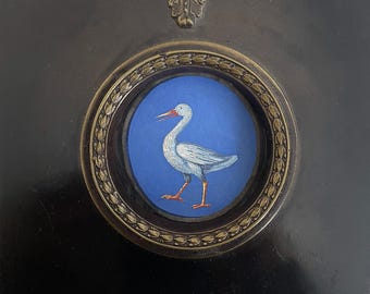 May include: A white bird with a long, red beak stands on a blue background. The bird is framed in a gold-colored, ornate frame.