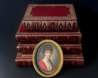 May include: An antique portrait in an oval gold frame rests in front of a stack of red leather-bound books with gold accents. The portrait depicts a woman in a red dress. The books have gold lettering and decorative borders.