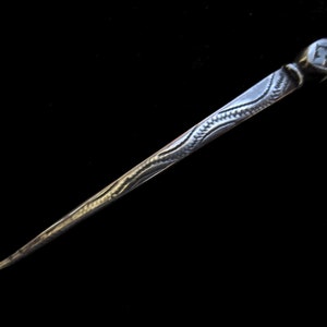 May include: A silver needle with a decorative design on the shaft. The needle has a pointed tip and a rounded head.