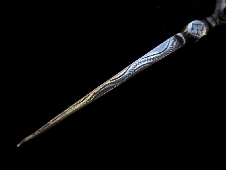 May include: A silver needle with a decorative design on the shaft. The needle has a pointed tip and a rounded head.