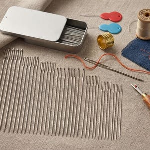 May include: A collection of silver-toned sewing needles arranged in rows, a rectangular metal storage tin, a gold-toned metal thimble, a wooden spool of thread, two small plastic needle threaders in red and blue, and a seam ripper with a wooden handle.