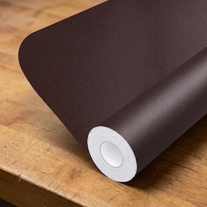 May include: A roll of dark brown adhesive contact paper with a textured, leather-like grain finish. The roll is partially unrolled, revealing a white paper backing on the inner core and a consistent, matte dark brown surface on the outer side.