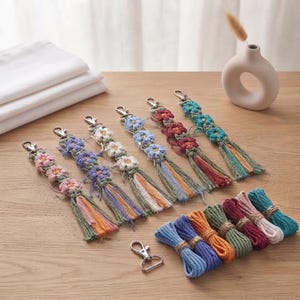 May include: Six macrame keychains featuring crocheted flower motifs in pink, purple, white, blue, red, and teal, each ending in a multi-colored tassel with a silver lobster clasp. A separate silver clasp and several bundles of colorful cotton cord are included.