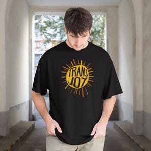 以下が含まれることがあります： 中央に「TRANS JOY」という文字がプリントされた、黄色の太陽のグラフィックが特徴の黒い半袖Tシャツ。太陽のデザインには、円形の中心から外側に向かって放射状の線が伸びており、大胆でコントラストの強い視覚効果を生み出しています。
