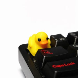 Pode incluir: Uma pequena figura de pato de borracha amarelo fixada em uma tecla de teclado de computador preto. O pato tem olhos pretos e um bico laranja. O teclado apresenta teclas pretas com texto vermelho, incluindo as palavras Tab e Caps Lock.