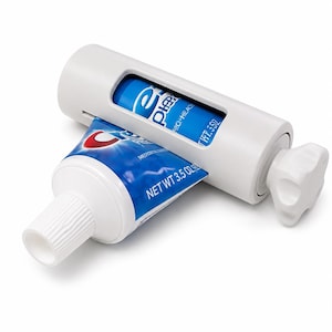 Peut inclure: Un presse-tube de dentifrice en plastique blanc doté d'un bouton rotatif manuel à une extrémité. L'appareil contient un tube de dentifrice Crest bleu et blanc d'un poids net de 99 grammes, partiellement inséré dans la fente centrale.