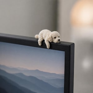 May include: A small, cream-coloured figurine of a curly-haired dog resting its front paws over the top edge of a black computer monitor. The dog has a textured coat, dark eyes, and a dark nose, positioned to look as if it is peering over the screen.