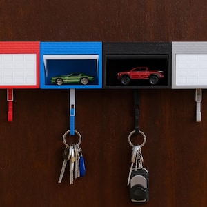 Peut inclure: Un ensemble de porte-clés muraux composé de quatre unités en forme de garage rouge, bleu, noir et gris. Chaque unité possède un crochet pour les clés. L'unité bleue contient une voiture miniature verte et l'unité noire un camion rouge. Deux unités ont des portes fermées.