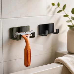 May include: An olive green wall-mounted razor holder with a vertical ribbed design, holding an orange razor with a silver blade. A second identical black wall-mounted holder is positioned to the right, featuring two hooks for storage.