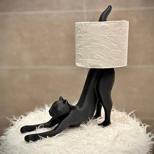 May include: A matte black cat-shaped toilet paper holder featuring a stylized feline in a stretching pose. The cat's back serves as a vertical spindle to hold a roll of white toilet paper with a subtle embossed floral pattern on its surface.