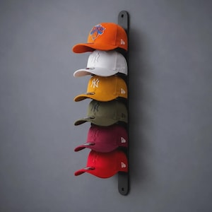 May include: A vertical black wall-mounted rack holding six baseball caps in orange, white, yellow, olive green, maroon, and red. Each cap features a New Era logo on the side and various team emblems on the front, organized in a single column.