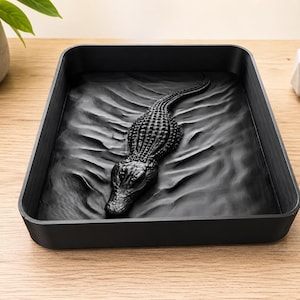 Könnte beinhalten: Ein rechteckiges schwarzes Tablett mit einem dreidimensionalen Relief eines im Wasser schwimmenden Alligators. Der Alligator ist in der Mitte positioniert, wobei sein strukturierter Rücken und Kopf über den modellierten Wellen des Bodens ragen.
