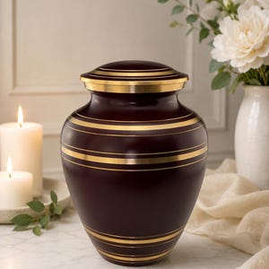 Puede incluir: Una urna funeraria de color burdeos intenso con una silueta redondeada y una tapa a juego. El recipiente está decorado con múltiples bandas horizontales de tono dorado que rodean el cuerpo y la tapa, creando un contraste con el acabado oscuro.