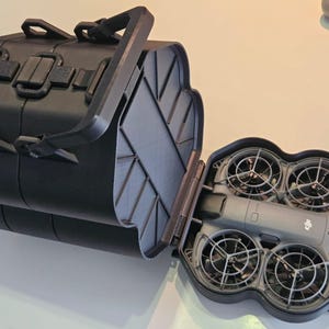 Op de afbeelding: Een zwarte harde draagkoffer met een handvat aan de bovenkant en veilige sluitingen, geopend om een grijze drone met vier beschermde propellers binnenin te onthullen. De drone heeft een compact ontwerp met een centrale behuizing.