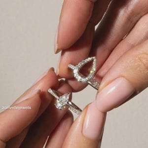 以下が含まれることがあります： 洋ナシ型のデザインが施された2つのシルバーカラーのリング。1つは中央に洋ナシ型の石が配置され、もう1つは小さな透明な石で縁取られた洋ナシ型のハローセッティングが特徴です。どちらのバンドも小さな透明な石でパヴェ装飾されています。テキストはzorivellejewelsと記載されています。