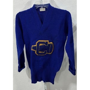 Peut inclure: Un pull à col en V et manches longues en tricot bleu roi. Le centre présente un grand écusson brodé jaune et bleu foncé représentant un porte-voix avec la lettre C à l'intérieur. Une étiquette blanche au col indique Jerry West Sporting Goods.