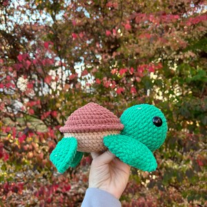 Peut inclure: Une tortue au crochet faite à la main dans les tons de vert, beige et rose. La tortue a une carapace et des nageoires texturées, avec un œil en bouton noir. La tortue est tenue devant un fond de feuillage d'automne coloré.