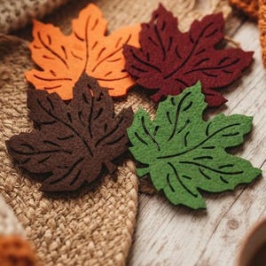 Puede incluir: Un juego de cuatro posavasos de fieltro con forma de hojas de arce en naranja, rojo oscuro, marrón oscuro y verde. Cada posavasos presenta detalles de nervaduras cortados con láser en la superficie, creando un patrón de hoja en el material.