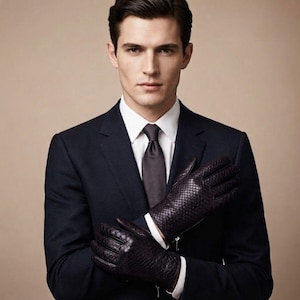 Peut inclure: Une paire de gants en cuir marron foncé avec un motif texturé rappelant la peau de serpent. Les gants présentent un petit détail de fermeture éclair argentée au poignet. Le matériau a une finition brillante et les gants sont portés sur une manchette de chemise blanche.