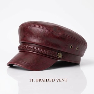 May include: A deep burgundy faux leather military-style cap featuring a structured crown and a short, curved visor. The hat is accented with a decorative braided band across the front, secured by two metallic buttons on the sides and metal eyelets.
