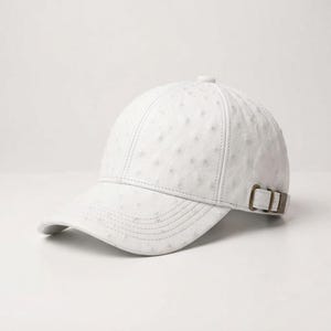May include: A white baseball cap featuring a textured ostrich-skin pattern across the crown and brim. The cap includes a curved visor with stitched detailing and an adjustable side strap secured with a metallic bronze-toned buckle hardware.