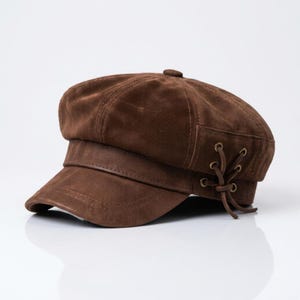 May include: A brown suede newsboy cap featuring a structured brim and a decorative side panel with brass-toned eyelets and a matching brown cord lace-up detail. The cap has a multi-paneled crown construction with a small button at the top center.