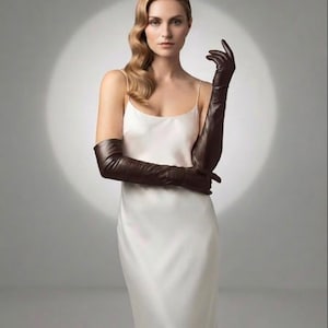 May include: A floor-length white satin slip dress with thin spaghetti straps, paired with long, dark brown leather opera-length gloves that extend past the elbows. The ensemble features a smooth, minimalist aesthetic with clean lines and a solid color palette.