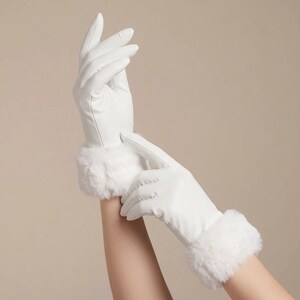 May include: A pair of white gloves featuring a smooth finish and a plush, white faux fur cuff at the wrist. The gloves are constructed with visible stitching along the fingers and palm, providing a clean, uniform appearance for the hands.