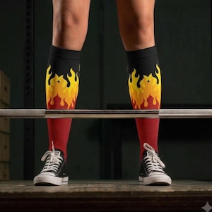 May include: A pair of knee-high athletic socks featuring a black upper section that transitions into a vibrant yellow, orange, and red flame pattern, ending in a solid red lower section, worn with black and white low-top canvas sneakers.