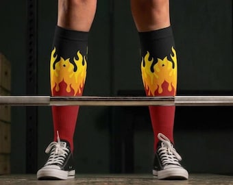 Puede incluir: Un par de calcetines deportivos hasta la rodilla con una sección superior negra que transiciona a un patrón de llamas vibrante en amarillo, naranja y rojo, terminando en una sección inferior roja sólida, usados con zapatillas de lona bajas.