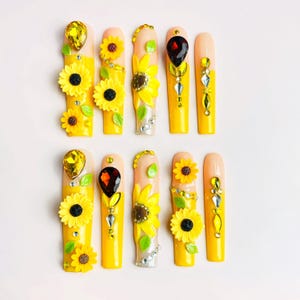 May include: A set of ten long, square-tipped press-on nails featuring a yellow and nude gradient base. The nails are decorated with three-dimensional yellow sunflower charms, green leaf accents, and various clear, yellow, and dark brown rhinestones.