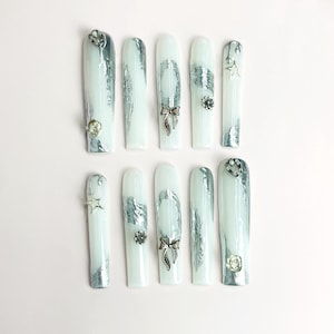 May include: A set of ten long, white press-on nails featuring metallic silver foil accents. The nails are decorated with small silver charms, including bows, stars, and flowers, arranged in various patterns across the glossy white surface.